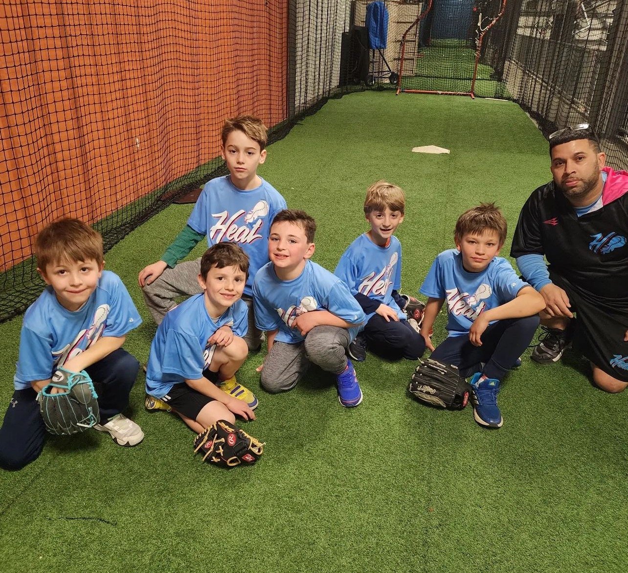 Group of young boys in blue baseball shirts posing with a coach indoors on artificial turf.