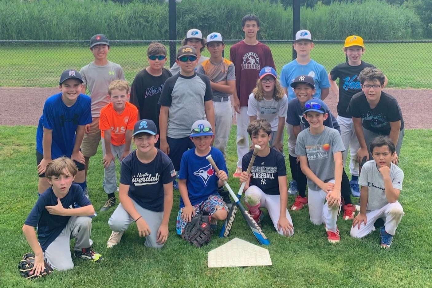 Group of kids posing on a baseball field, some wearing sports caps and shirts, two holding baseball bats, with a home plate on the grass.